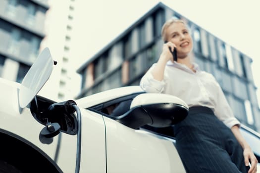 Focus charging-electric car with EV charger at charging station with blur businesswoman talking on phone with residential building in background as progressive lifestyle concept.