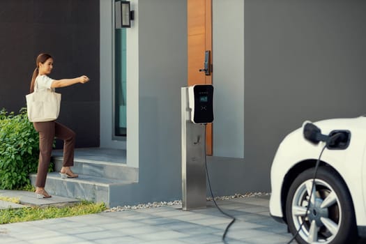 Asian woman lock electric vehicle with remote car key at her garage, EV car recharge battery at home charging station. Alternative clean energy applied in daily life as progressive lifestyle concept.