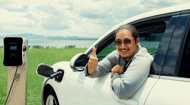 Progressive man connect power cable plug from charging station to electric car, EV car driven by green energy on green field for future clean environment with beautiful cloudscape background concept.