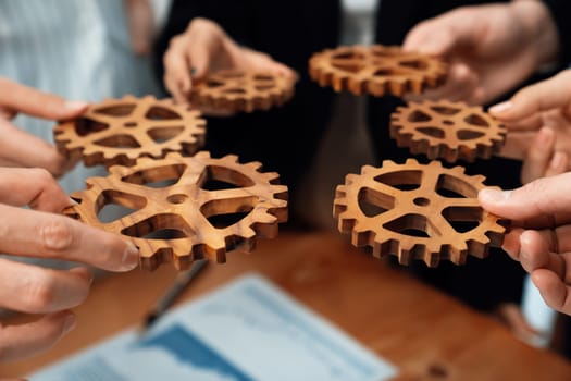 Closeup hand holding wooden gear by businesspeople wearing suit for harmony synergy in office workplace concept. Group of people hand making chain of gears into collective form for unity symbol.