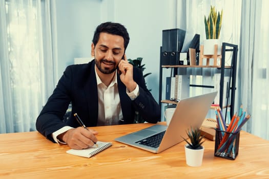 Diligent businessman busy talking on the phone call with clients while working with laptop in his office desk as concept of modern hardworking office worker lifestyle with mobile phone. Fervent