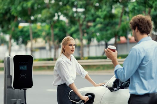 Progressive businessman and businesswoman with electric car parking and connected to public charging station before driving around city center. Eco friendly rechargeable car powered by clean energy.