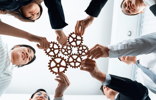 Hand holding wooden gear by businesspeople wearing suit for harmony synergy in office workplace. Bottom view office worker hand make chain of gear into collective unity form.