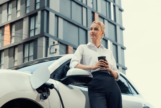 Businesswoman drinking coffee, leaning on electric vehicle recharging at public charging station with residential apartment condos building in background as progressive lifestyle by eco-friendly car.