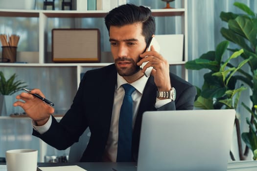 Diligent businessman busy talking on the phone call with clients while working with laptop in his office as concept of modern hardworking office worker lifestyle with mobile phone. Fervent