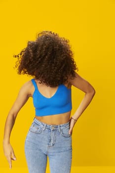 Woman with curly afro hair in a blue T-shirt on a yellow background dancing flying hair with sunglasses yellow, hand signs, look into the camera, smile with teeth and happiness, copy space. High quality photo