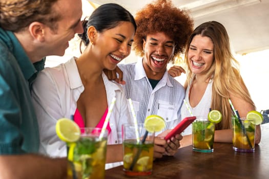 Group of happy multiracial friends looking at mobile phone in a beach bar having mojito cocktails. Technology and leisure activity.