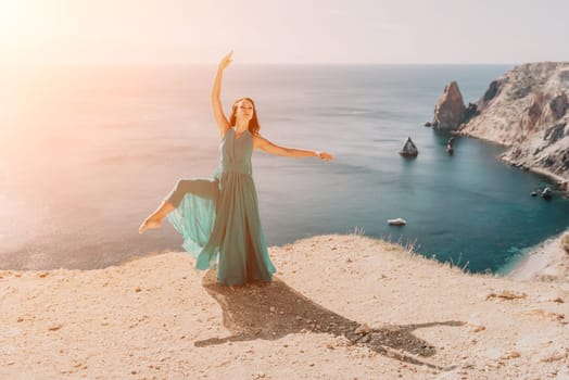 Side view a Young beautiful sensual woman in a red long dress posing on a rock high above the sea during sunrise. Girl on the nature on blue sky background. Fashion photo.