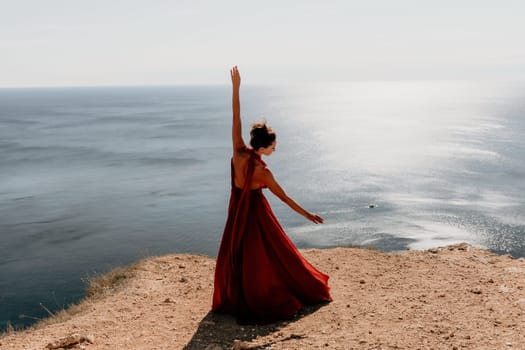 Side view a Young beautiful sensual woman in a red long dress posing on a rock high above the sea during sunrise. Girl on the nature on blue sky background. Fashion photo.