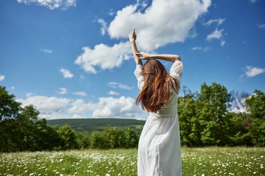 a woman stands with her back to the camera in a light dress in a chamomile field with her hands raised above her head. High quality photo