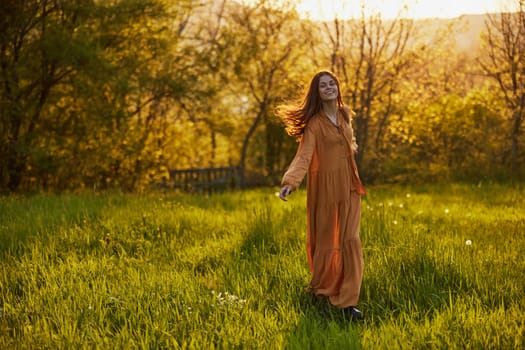 a joyful woman stands green in a field smiling at the camera, in a long orange dress, illuminated by the warm rays of the setting sun. High quality photo