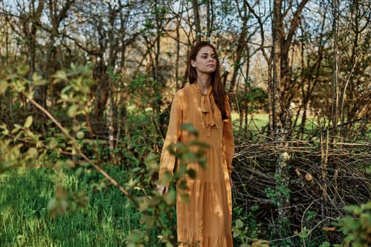 a beautiful, slender woman with long hair walks in the shade near the trees, dressed in a long orange dress, enjoying the weather and the weekend. The theme of privacy with nature. High quality photo