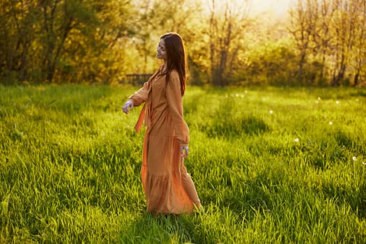 a relaxed slender woman enjoys the sunset standing in a green field with tall grass in an orange dress posing looking away. High quality photo