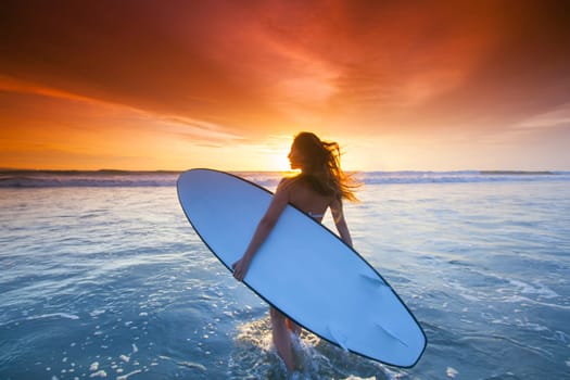 A beautiful young slim sporty woman in bikini with a surfboard is standing at ocean beach at sunset