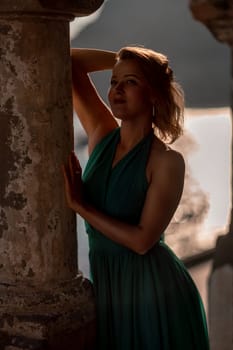 Happy blonde in a long mint dress posing against the backdrop of the sea in an old building with columns. Girl in nature against the blue sky