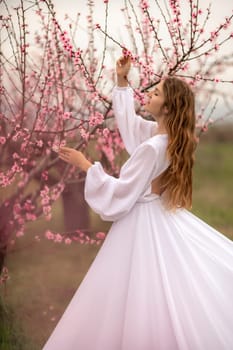 Woman peach blossom. Happy curly woman in white dress walking in the garden of blossoming peach trees in spring.