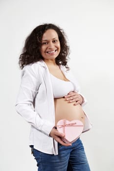 Happy multi ethnic pregnant woman, future mom caressing her belly, smiles at camera, shows a cute pink heart shaped gift box with happy present for Mother's or Women's Day, isolated white background
