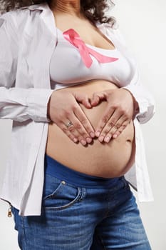 Closeup of pregnant woman wearing pink awareness ribbon, putting hands on her belly, making heart shape, isolated on white background. Breast Cancer Awareness campaign. Gynecology and women's health