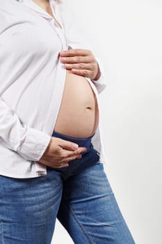 Midsection of a pregnant woman, expecting baby, enjoying her 28 week of pregnancy, holding hands on her bare belly, isolated on white background. Close-up of gravid female. Maternity. Motherhood.