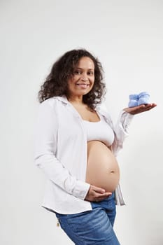 Excited positive multi ethnic pregnant woman wearing unbuttoned shirt and blue jeans, holding blue knitted baby booties, caressing belly, smiling cutely looking at camera, isolated on white background