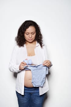 Pregnant curly haired woman in second semester of pregnancy, holding blue baby bodysuit, preparing to child birth. Young gravid mother with big belly waiting of a baby, isolated on white background