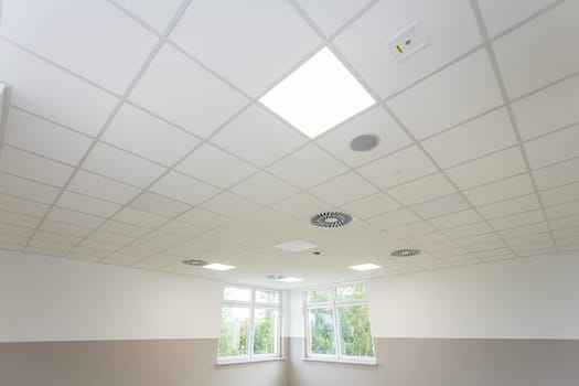 Suspended panel ceiling seen from below with lamps lit. Just below the ceiling are two windows.