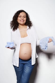 Cheerful happy curly haired pregnant woman holding cute blue baby booties and bodysuit for her future newborn child, smiling at camera, isolated on white background. Maternity. Pregnancy 2nd trimester