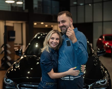 Happy caucasian couple hugging and man holding new car keys in car showroom