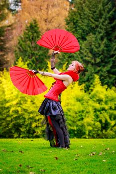 Young beautiful woman in circus costume play with red fans in the park.