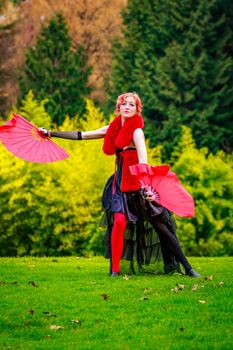 Young beautiful woman in circus costume play with red fans in the park.