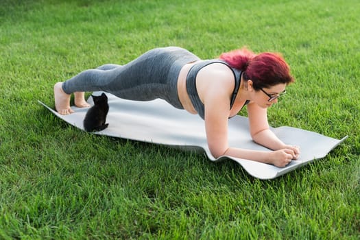 Young plus size woman in sporty top and leggings standing in plank on yoga mat spending time on green grass in yard. Black kitten walks around her. Well being fitness concept