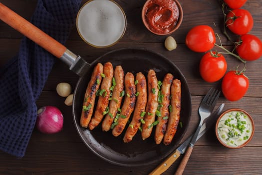 Delicious sausages cooked in a frying pan with vegetables, various sauces and a glass of beer on a wooden table.