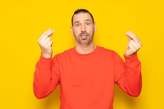 Bearded Hispanic man in his 40s wearing a red jumper making the Italian mamma mia gesture isolated on yellow studio background