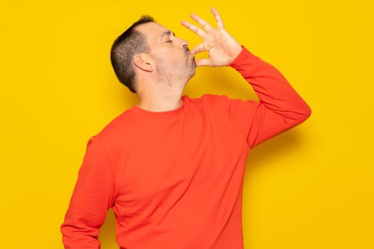 Bearded Hispanic man in his 40s wearing a red jumper smacking his lips and licking his fingers after devouring a delectable delicacy, isolated against a yellow studio