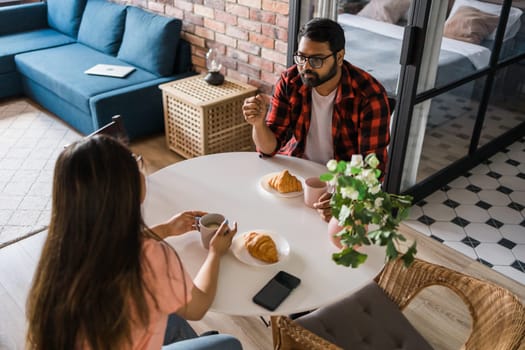 Young diverse loving couple eating croissant and talks together at home in breakfast time. Communication and relationship