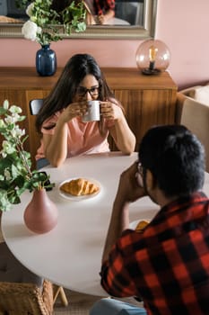 Happy couple eating breakfast and talking at dining table in morning. Indian girl and latin guy. Relationship and diversity