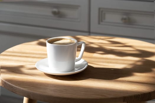 A white porcelain cup with coffee on a saucer stands on a small round table made of natural wood. Morning, coffee, hot drink. Hard light.