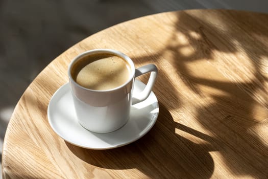 A white porcelain cup with coffee on a saucer stands on a small round table made of natural wood. Morning, coffee, hot drink. Hard light.