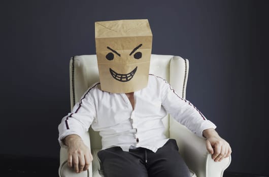 A man with a paper bag on his head, with a drawn angry smiley face, sits in a white chair. Emotions and gestures.