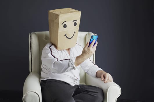 A man in a white shirt with a paper bag on his head, with a drawn smiling emoticon, is talking on a video call on a smartphone, gesturing with his hand.