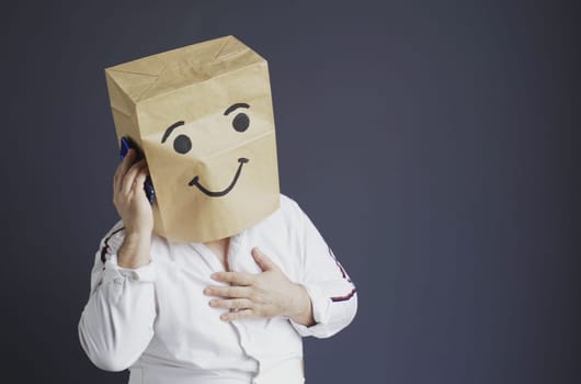A man in a white shirt with a paper bag on his head, with a smiley face drawn, talking on the phone.