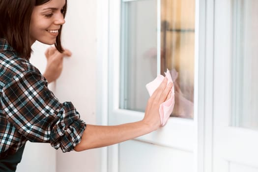 A young girl in an apron does housework, washes the windows with a special product, wipes it dry with a rag, a woman washes the glass with a sponge.