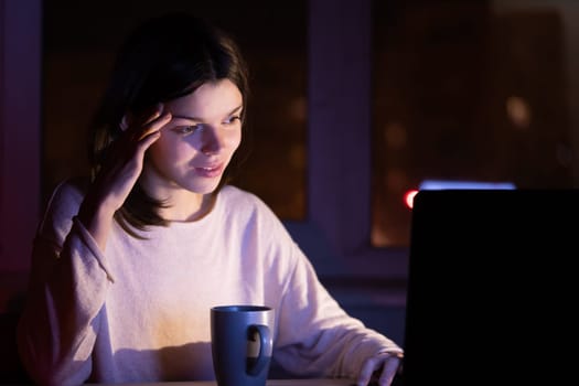 Young girl works at a laptop online at night against the background with the city lights outside the window. Woman is drinking coffee, holding a cup in her hands and reading the text on her device.