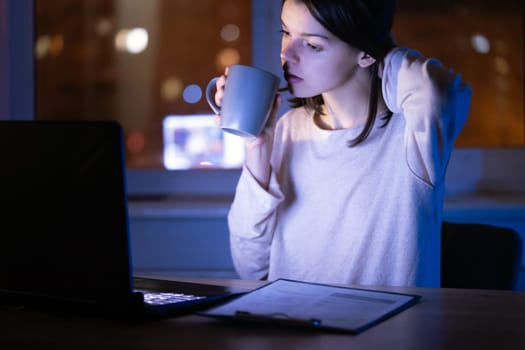 Young girl works at a laptop online at night against the background with the city lights outside the window. Woman is drinking coffee, holding a cup in her hands and reading the text on her device.