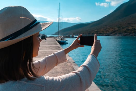 A young girl in a hat and with a backpack makes a photo of a beautiful bay with yachts and boats on the sea while traveling, a tourist walks along the shore next to the ocean and takes pictures.