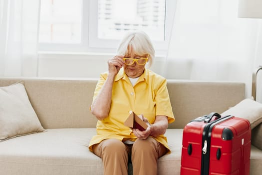 Happy senior woman with passport and travel ticket packed a red suitcase, vacation and health care. Smiling old woman joyfully sitting on the sofa before the trip raised her hands up in joy. High quality photo