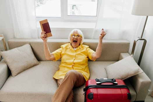 Happy senior woman with passport and travel ticket packed a red suitcase, vacation and health care. Smiling old woman joyfully sitting on the sofa before the trip raised her hands up in joy. High quality photo