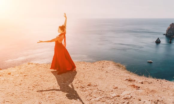 Side view a Young beautiful sensual woman in a red long dress posing on a rock high above the sea during sunrise. Girl on the nature on blue sky background. Fashion photo.