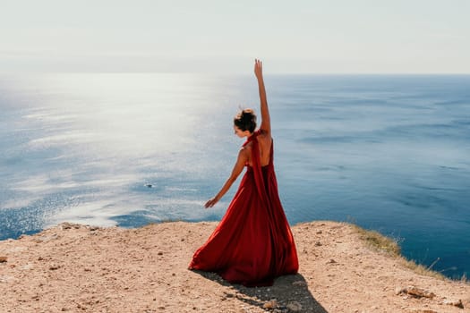 Side view a Young beautiful sensual woman in a red long dress posing on a rock high above the sea during sunrise. Girl on the nature on blue sky background. Fashion photo.