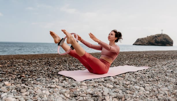 Middle aged well looking woman with black hair doing Pilates with the ring on the yoga mat near the sea on the pebble beach. Female fitness yoga concept. Healthy lifestyle, harmony and meditation.